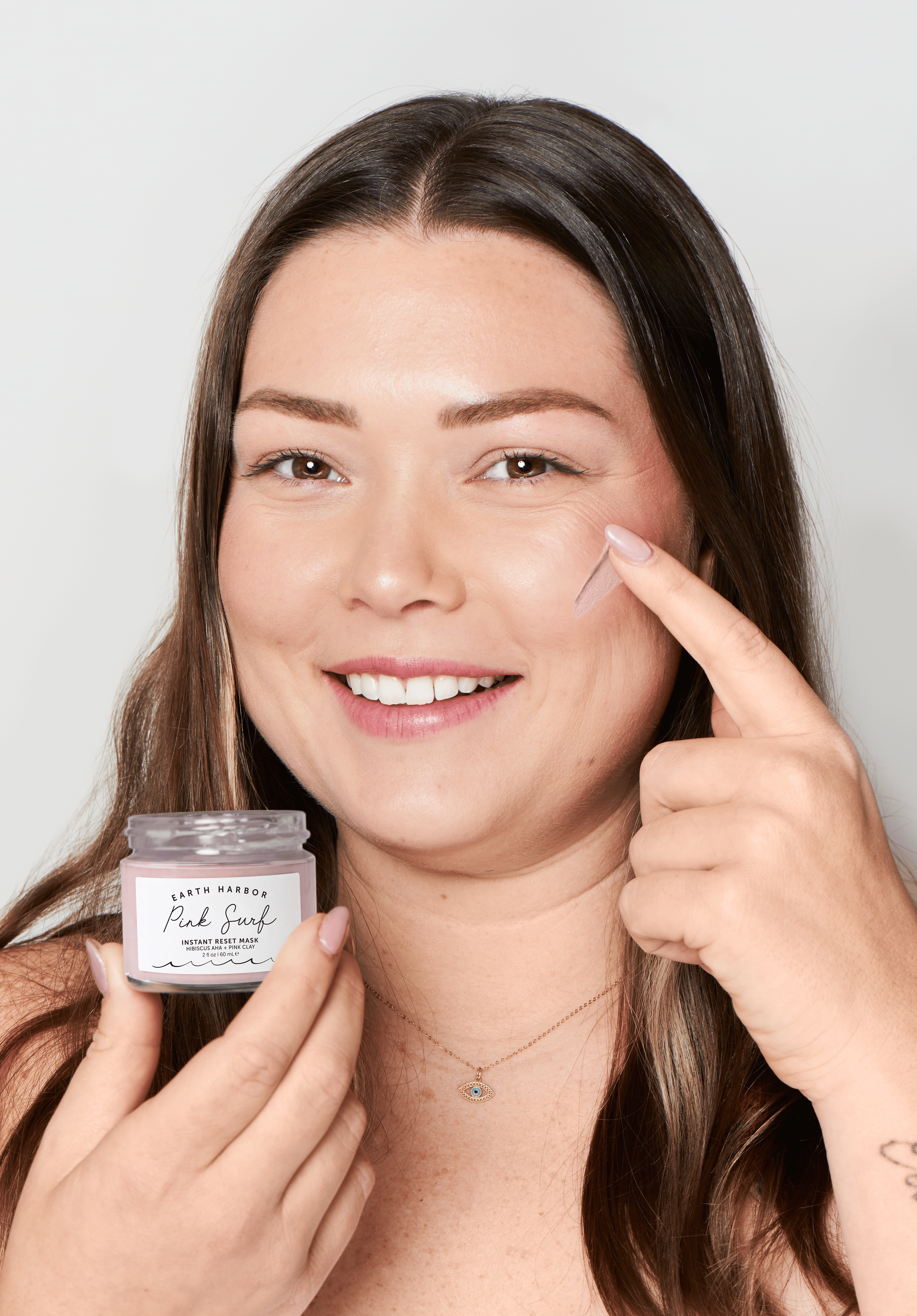 Woman applying mask to her face with a jar of mask in her hand on a plain background